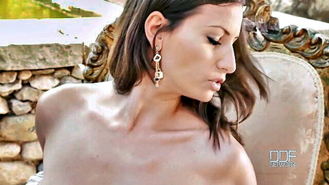 Sensual topless brunette with dangling earrings poses sideways on ornate outdoor chair