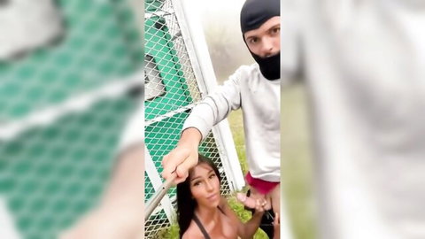 Masked man leashes busty transsexual at fence outdoors