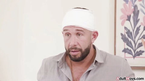 Bearded man with white head bandage sits talking in bedroom wearing open shirt