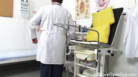 Male doctor in white lab coat stands by yellow gyno exam chair in mature medical office