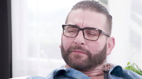 Mature bearded man with glasses and neck tattoo looking intense in blue shirt living room