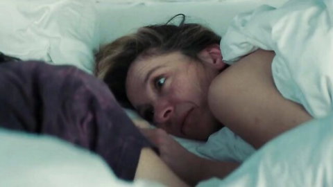 Blonde milf lying naked under white sheets with bare shoulder and intense gaze