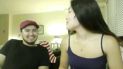 Smiling Asian woman in tight tank top flirts with capped guy on couch