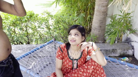 Smiling South Asian beauty in red saree sits on hammock with shirtless man outdoors teasing