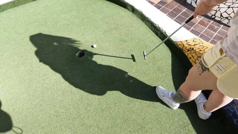 Tattooed babe in tight yellow shorts bends over showing big ass putting golf ball on outdoor mini golf course