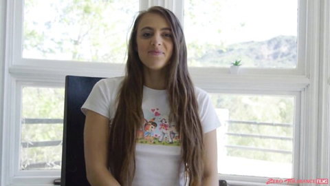 Gia Derza in white t-shirt with cartoon print, long hair, on sofa for domination scene