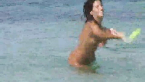 Nude woman smiling in shallow beach water, holding yellow object, amateur nudist