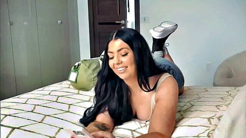 Seductive brunette MILF with big tits and thick ass in tight tank top and shorts teases smiling on bed
