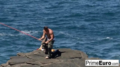 Muscular shirtless hunk kneeling on rocks pulling red rope from ocean waves at beach
