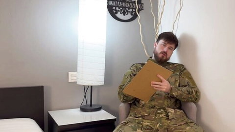 Bearded military man in camo uniform sits holding folder in bedroom