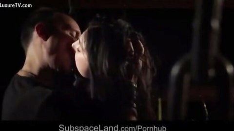Dominant man passionately kisses submissive brunette slave in dark BDSM dungeon.