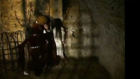 Auburn midwife in white dress interrogated by cloaked figure in dim dungeon
