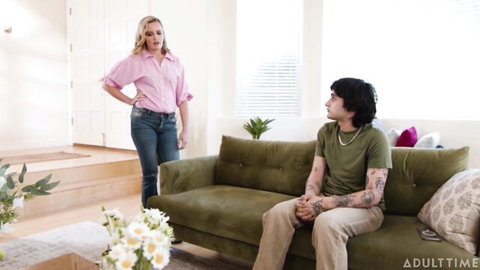 Blonde MILF in pink shirt and jeans confronts tattooed guy sitting on green sofa in taboo living room scene