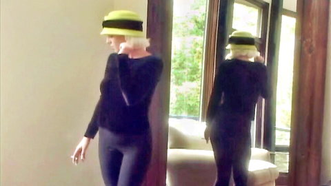 Sexy blonde babe in tight black leggings and yellow hat poses teasingly in mirror smoking cigarette
