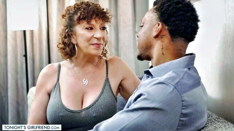 Mature brunette MILF with huge tits in gray tank top gazing seductively at black stud on couch