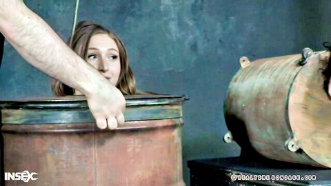 Man grips young brunette slave's chin hard while her head is trapped in rusty metal barrel BDSM bondage