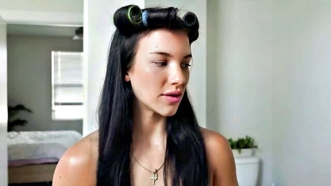 Brunette woman with long black hair in blue rollers, bare shoulders, thoughtful expression