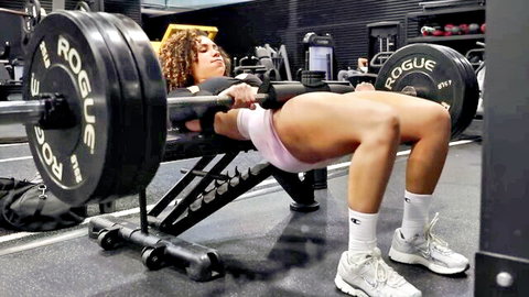 Curly-haired Black woman hip thrusting barbell for juicy glutes in pink shorts