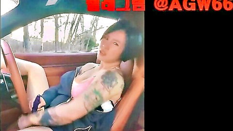 Tattooed Asian woman in unzipped pink top showing cleavage in car