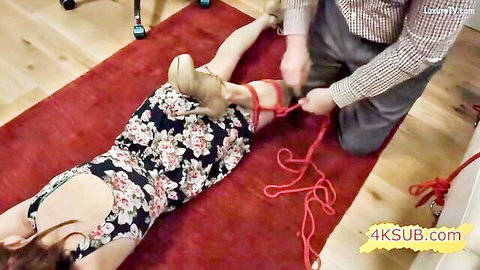 Redhead girl in floral dress bound with tight red ropes, man tying her legs