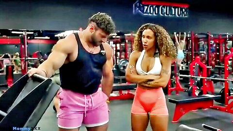 Muscular hunk in pink shorts holds gym mat eyeing busty curly-haired babe in tight white sports bra and shorts