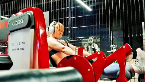 Blonde babe in tight jeans and sports bra doing leg curls on Cybex Eagle NX gym machine