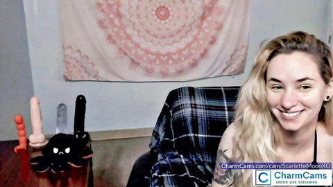 Smiling blonde webcam babe Scarlette Moon teases with colorful dildos on table in plaid shirt