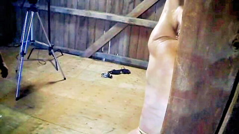 Nude slim blonde woman in bondage bent over wooden beam in brutal BDSM scene