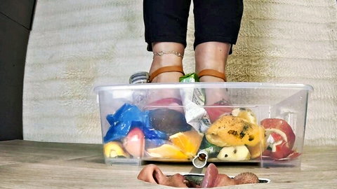 Woman in black pants and strappy heels stepping into fruit-filled trash bin for fetish play