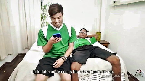 Two athletic guys in green soccer jerseys and shorts on bed, one on phone