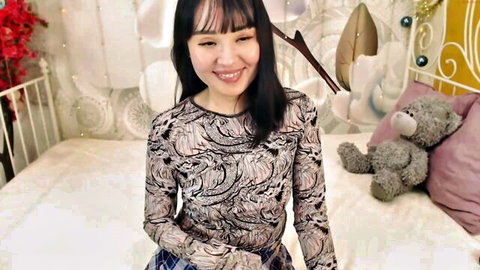 Smiling Asian babe in sheer patterned top and plaid skirt posing on cute teddy bear bed webcam