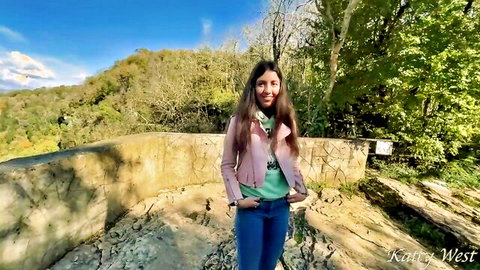 Katie West smiling in pink jacket and jeans outdoors in woods, public pickup tease