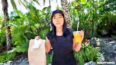 Emerald Loves in black dress holding delivery bag and orange drink outdoors