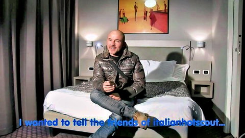 Bald man in shiny jacket sits smiling on hotel bed for Italian hotscout casting intro