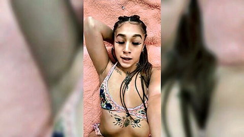 Tattooed mixed-race woman in colorful bikini top, braided hair, solo selfie on pink surface