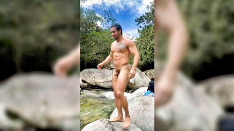 Muscular tattooed hunk stands nude with huge erect cock on river rocks outdoors