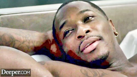 Tattooed black stud lounging shirtless with open mouth on bed