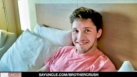 Cute young guy in pink shirt smiling on bed for BrotherCrush gay taboo preview