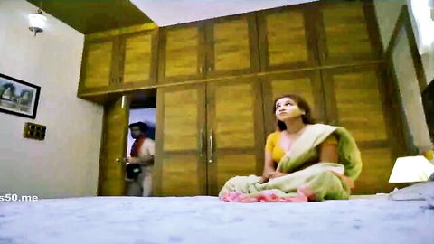 Desi woman in sheer yellow saree with deep cleavage sits on bed, man peeks from door in Ullu series