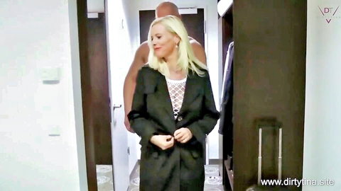 Seductive blonde in black coat smiles back at muscular black stud standing close behind her in hotel room