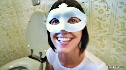 Smiling masked woman in white t-shirt poses beside toilet in bathroom for slave humiliation