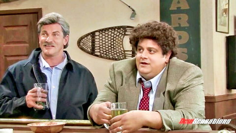 Silver-haired mustached man and curly-haired chubby guy in suit chatting at wooden bar holding drinks
