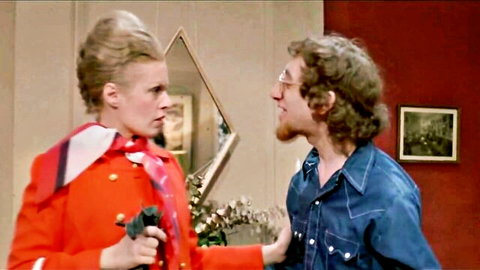 Blonde woman in red uniform holds gun at man in tense roleplay porno scene