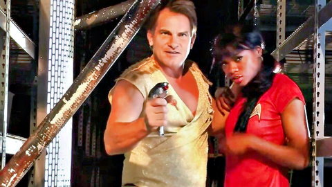 Muscular man in gold Star Trek uniform holds phaser on sexy ebony woman in red uniform parody scene