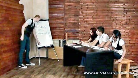 Male teacher in vest points at whiteboard while clothed brunette students watch in CFNM femdom classroom