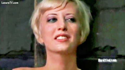 Smiling blonde submissive with heavy makeup in dark BDSM dungeon