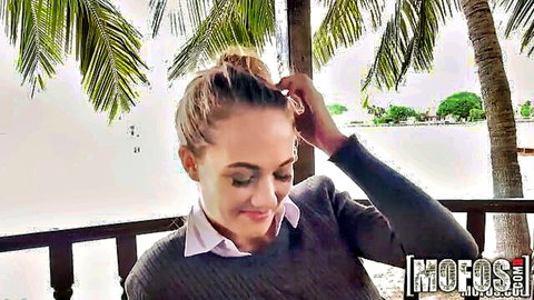 Blonde woman in gray sweater smiling seductively outdoors at tropical beach resort