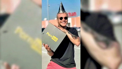 Tattooed guy in sunglasses and party hat smiling while holding large Pornhub box outdoors