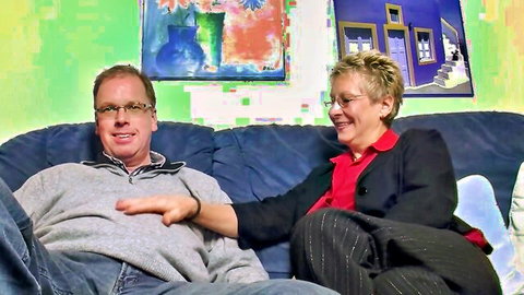 Mature glasses-wearing couple smiling intimately on couch with hand on thigh
