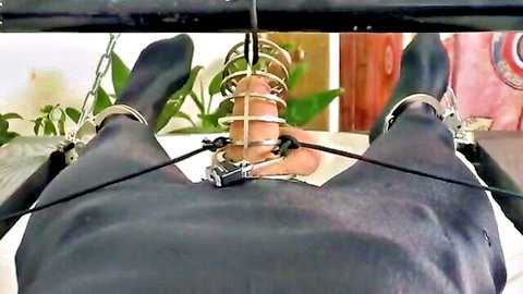 Man in metal chastity cage, legs chained and spread in BDSM restraint